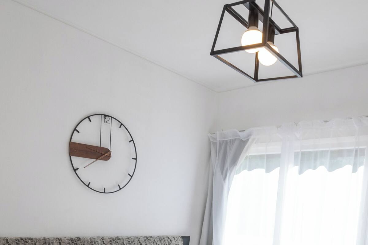 A neatly made bed with white linens, a round wall clock, and a geometric pendant light.