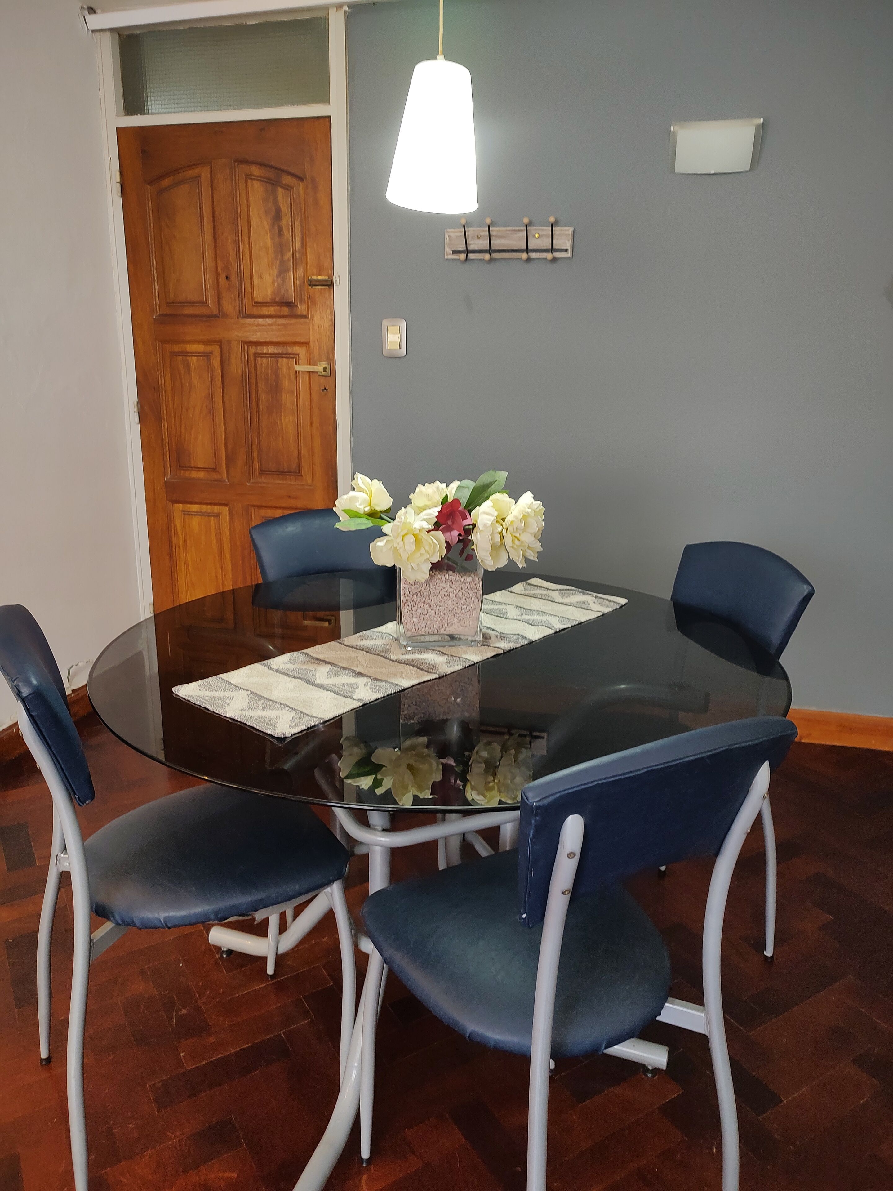 Una mesa redonda de vidrio con un posavasos blanco, rodeada por cuatro sillas azules con marcos blancos. Un jarrón con flores blancas y rosas está sobre la mesa.