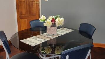 Una mesa redonda de vidrio con un posavasos blanco, rodeada por cuatro sillas azules con marcos blancos. Un jarrón con flores blancas y rosas está sobre la mesa.