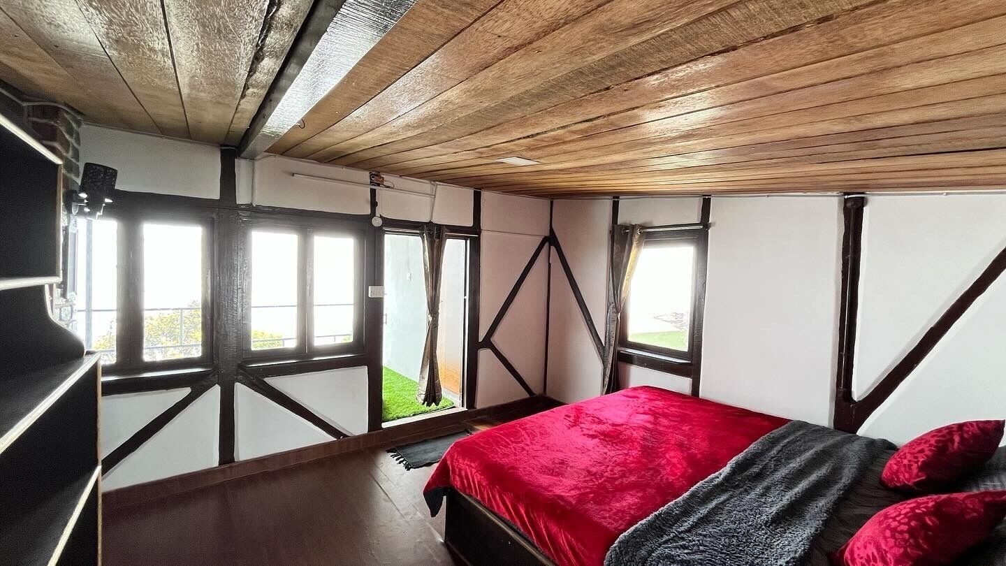 A bedroom with a red bedspread, a wooden ceiling, and large windows.