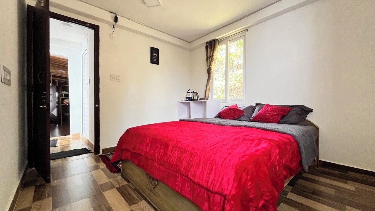 A bedroom with a red bedspread, a grey blanket, and a window with curtains.