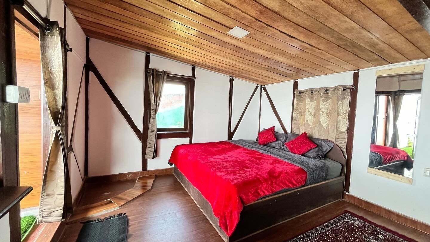 A bedroom with a wooden ceiling, a bed with red and grey bedding, and a window with curtains.