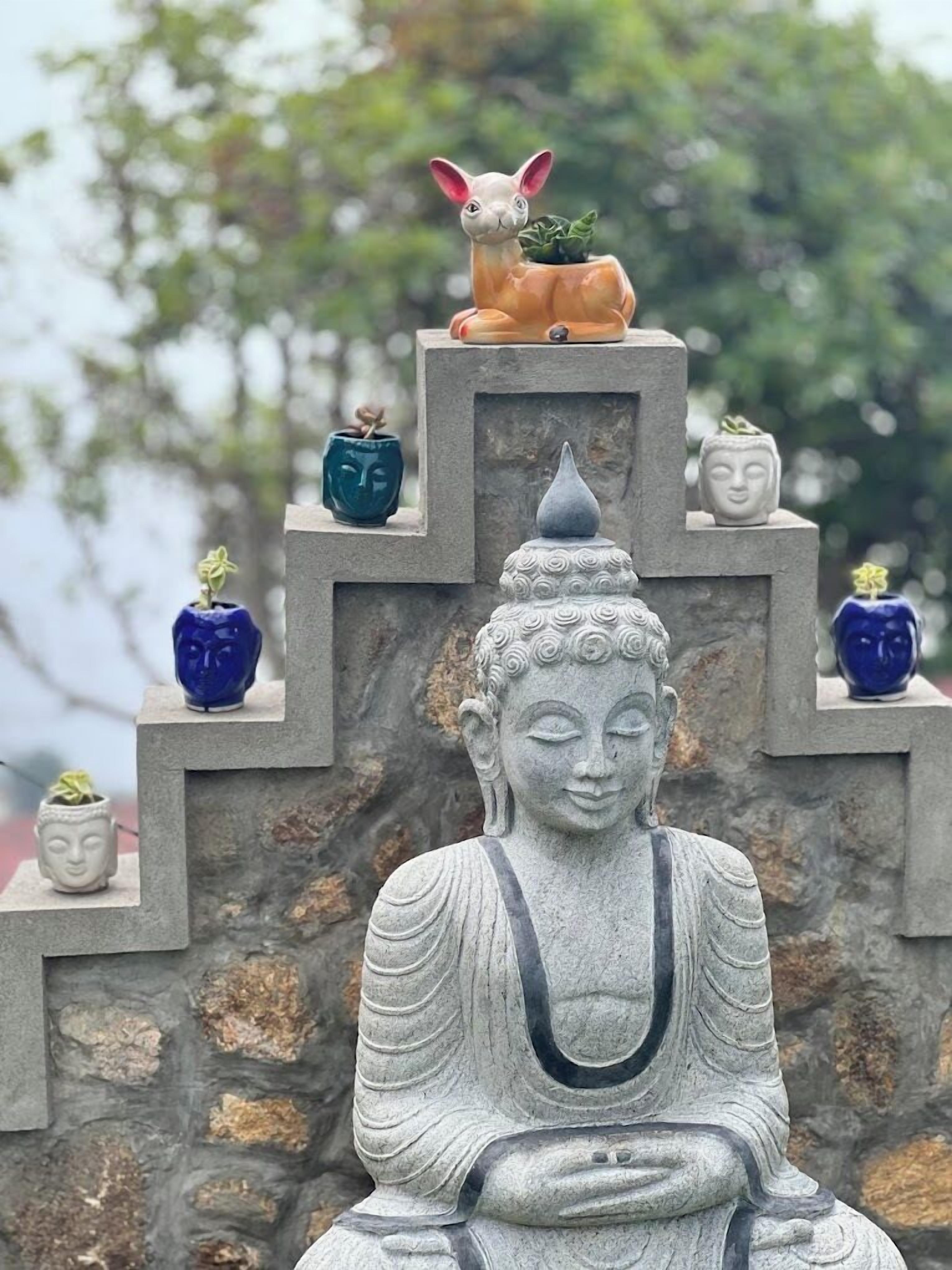 A stone Buddha statue with a seated figure and a smaller figure on top, surrounded by various small statues and plants on a stone structure.