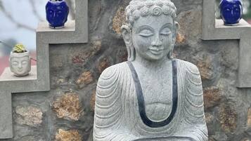 A stone Buddha statue with a seated figure and a smaller figure on top, surrounded by various small statues and plants on a stone structure.