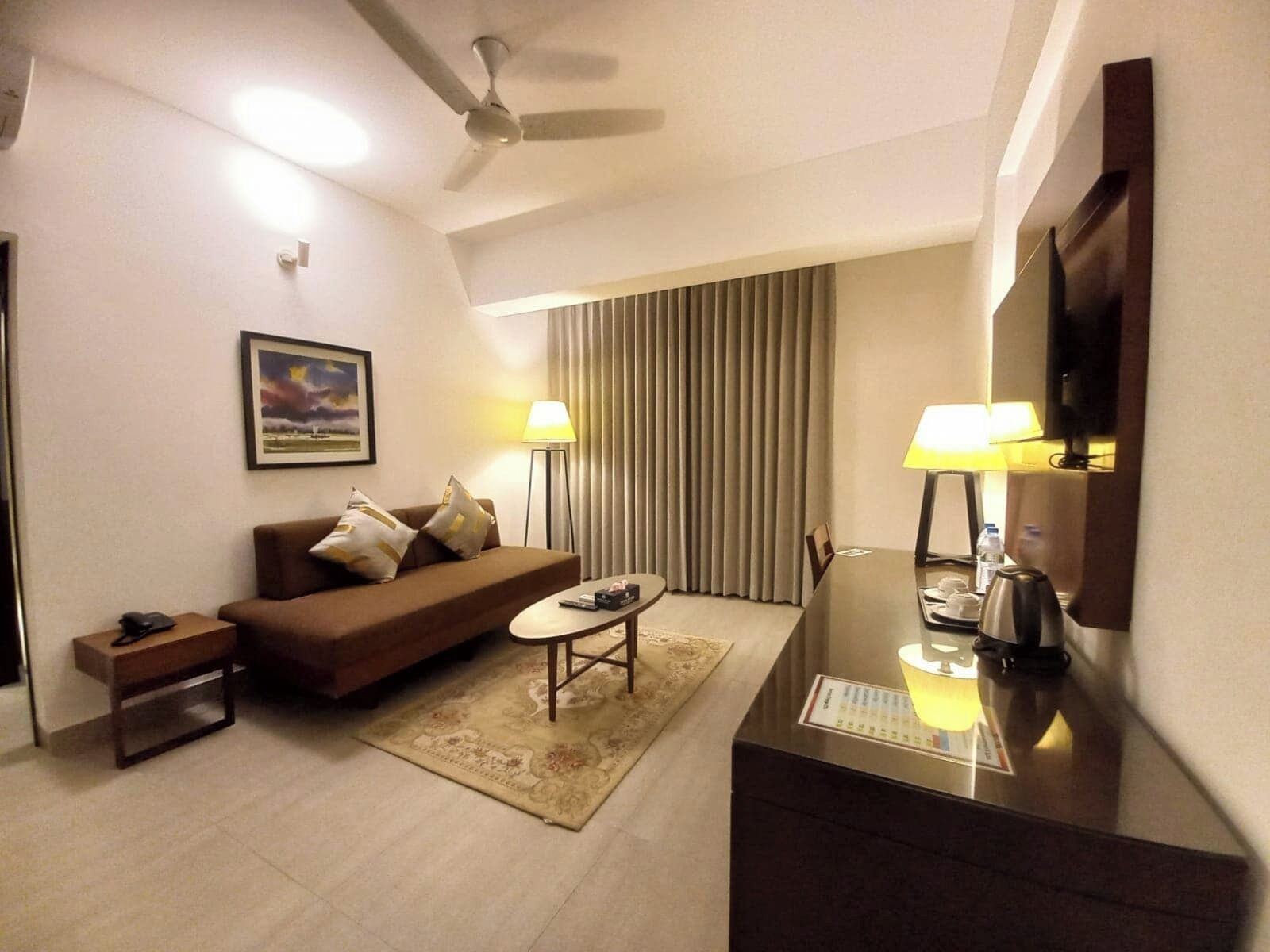 Una habitación de hotel moderna con un sofá marrón, una pequeña mesa de centro, un escritorio con lámpara, un ventilador de techo y un ventanal con cortinas.