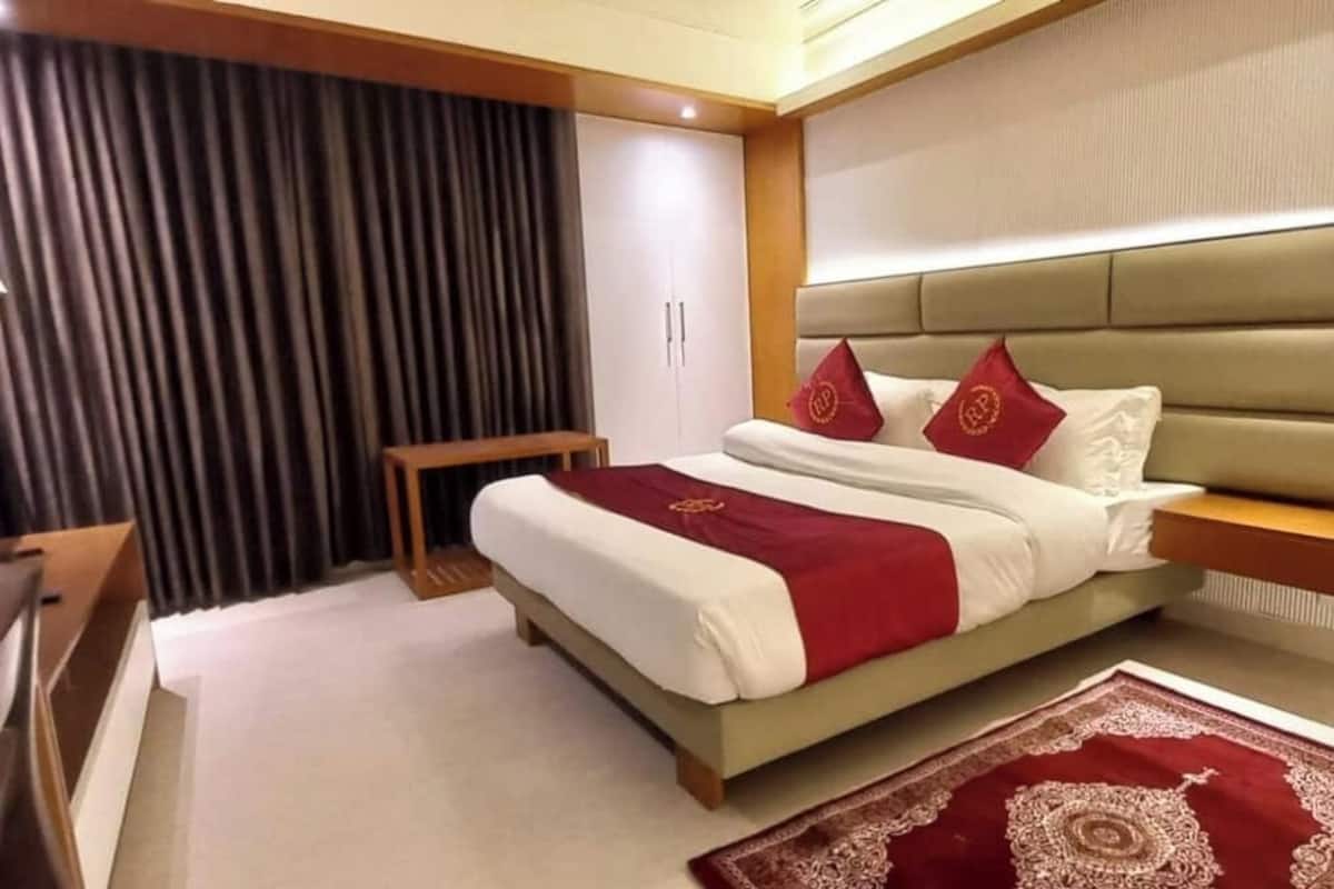 A hotel room with a bed, a bedside table, a lamp, a dark curtain, and a red patterned rug.