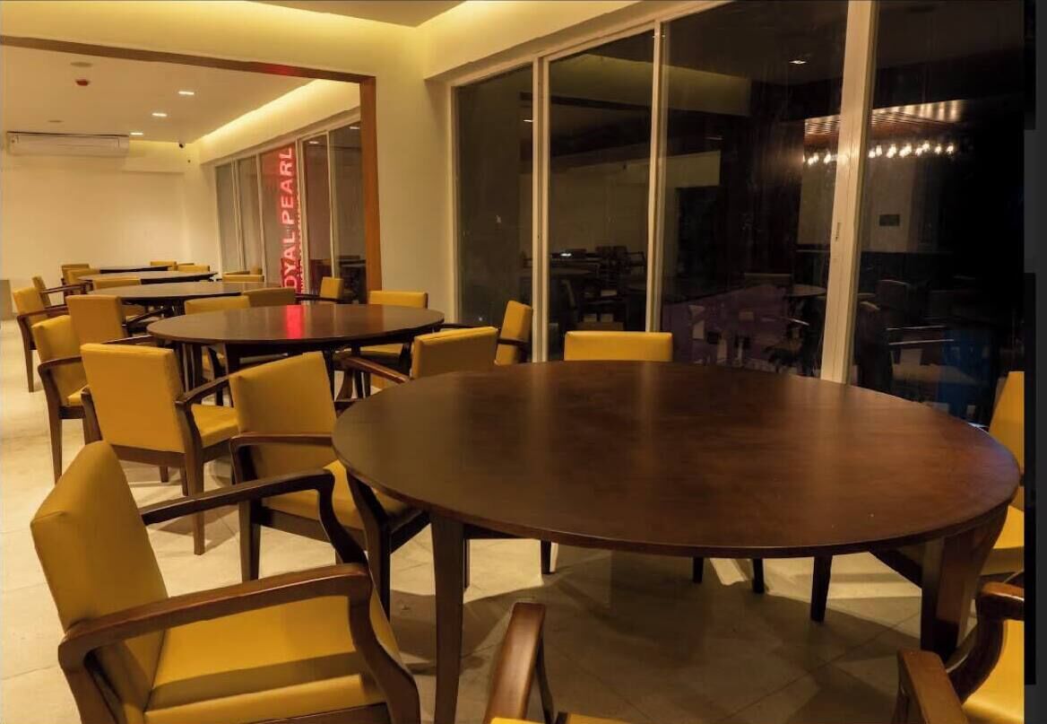 A conference room with a large oval table surrounded by yellow chairs, large windows, and a visible air conditioning unit.