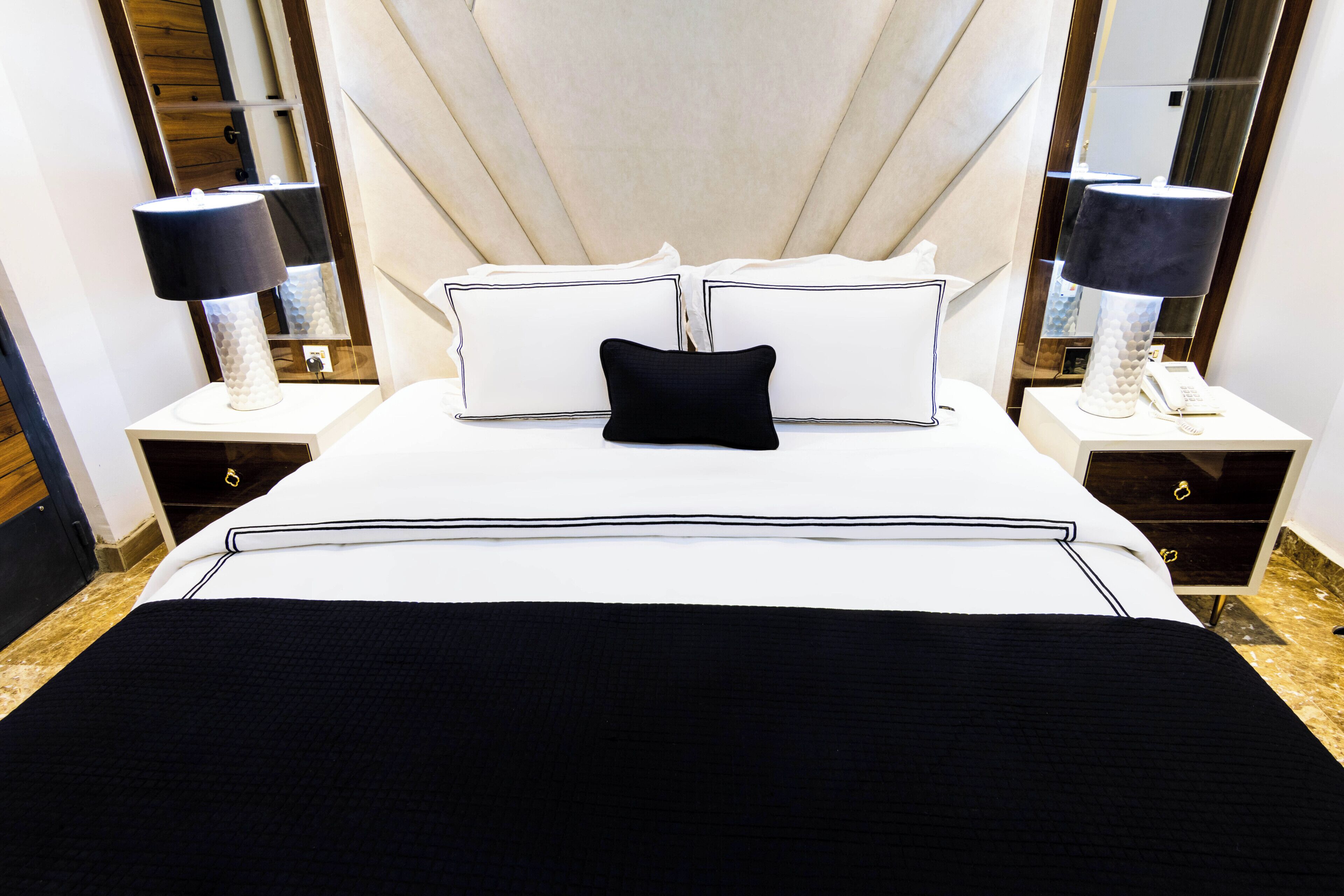 Un lit bien fait, avec une literie blanche et noire, deux tables de chevet avec des lampes et un grand miroir au-dessus du lit.