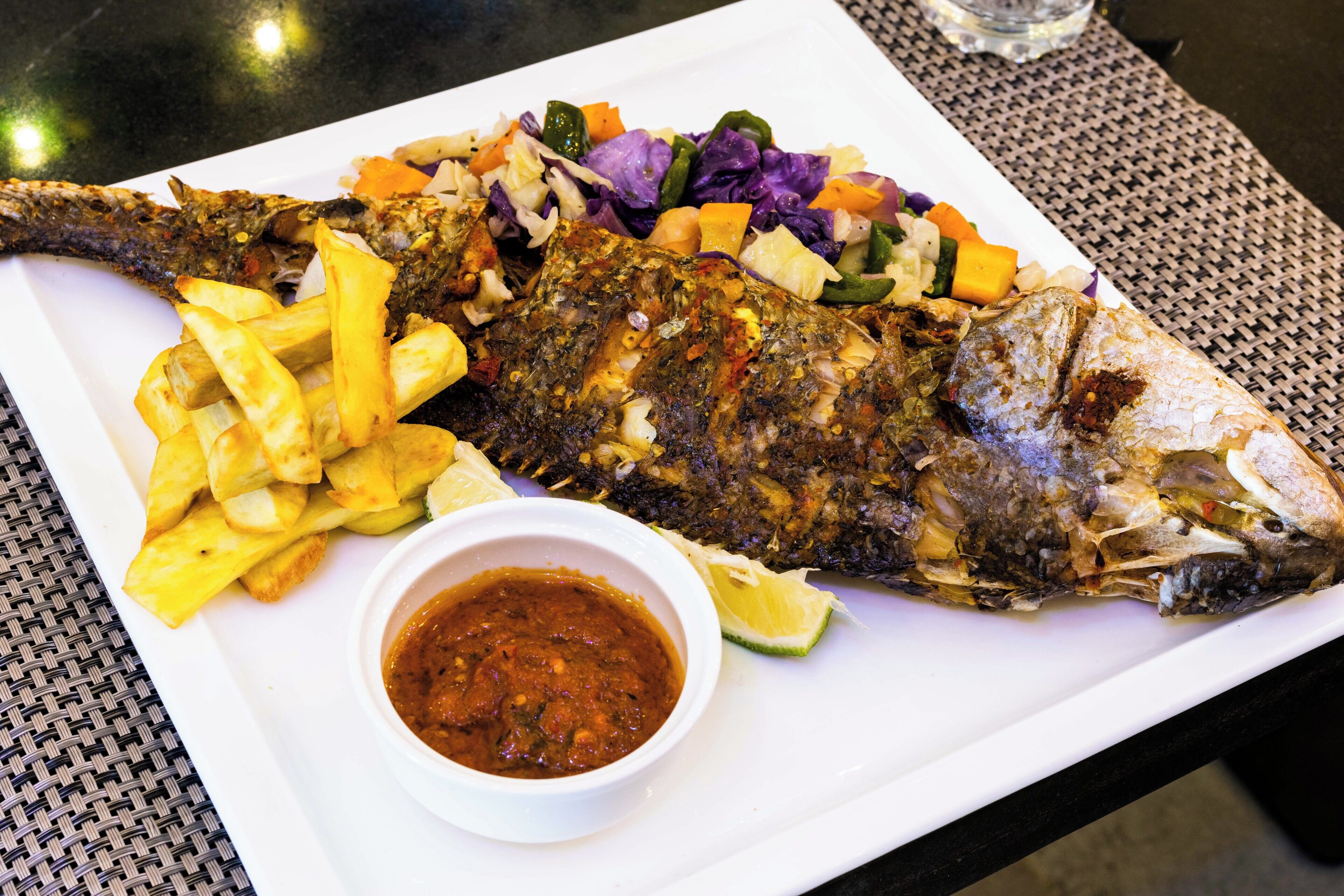 A white plate with a whole grilled fish, French fries, a side salad, and a bowl of sauce.