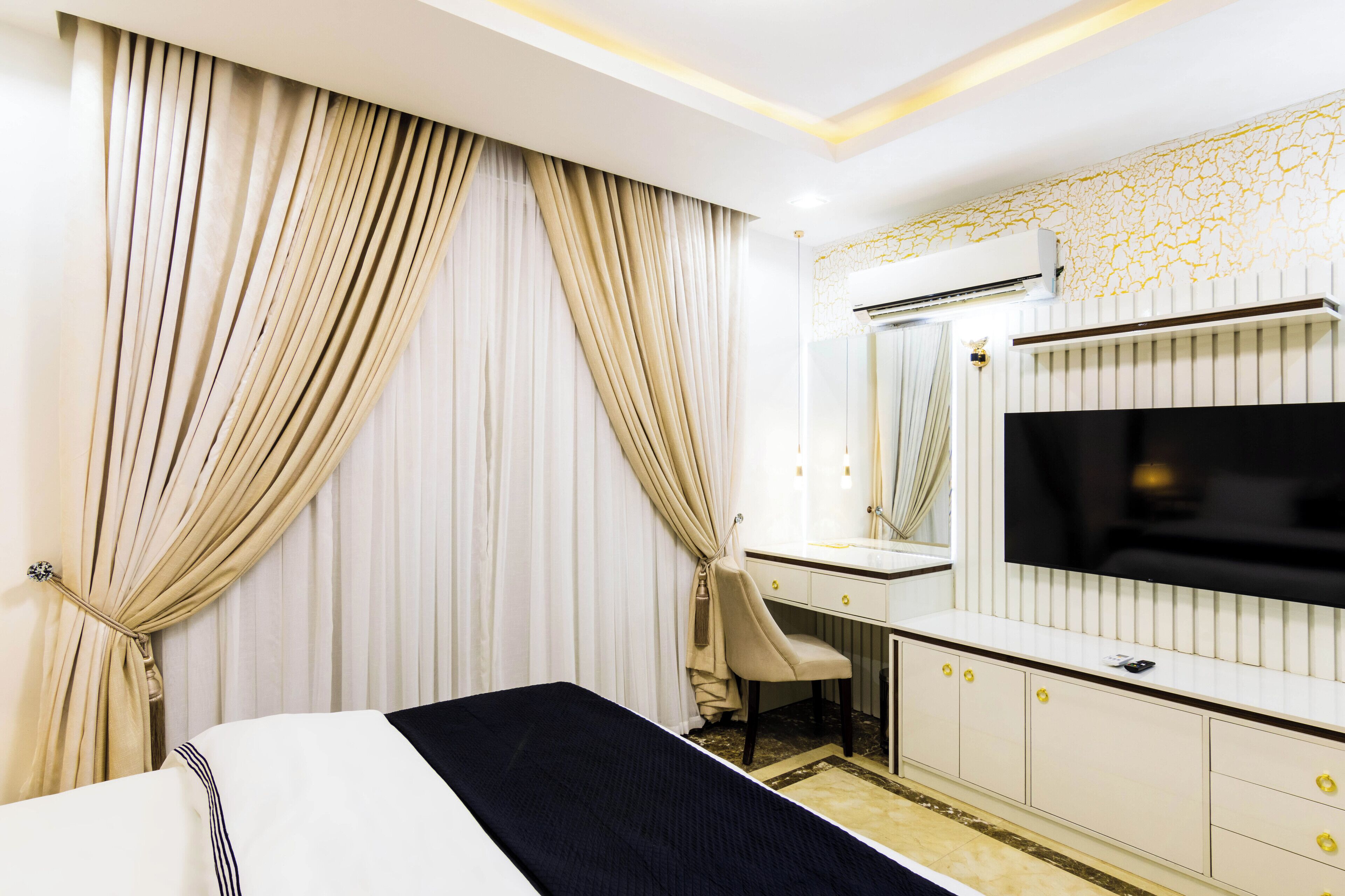 A hotel room with a bed, a desk with a chair, a TV, and an air conditioning unit.