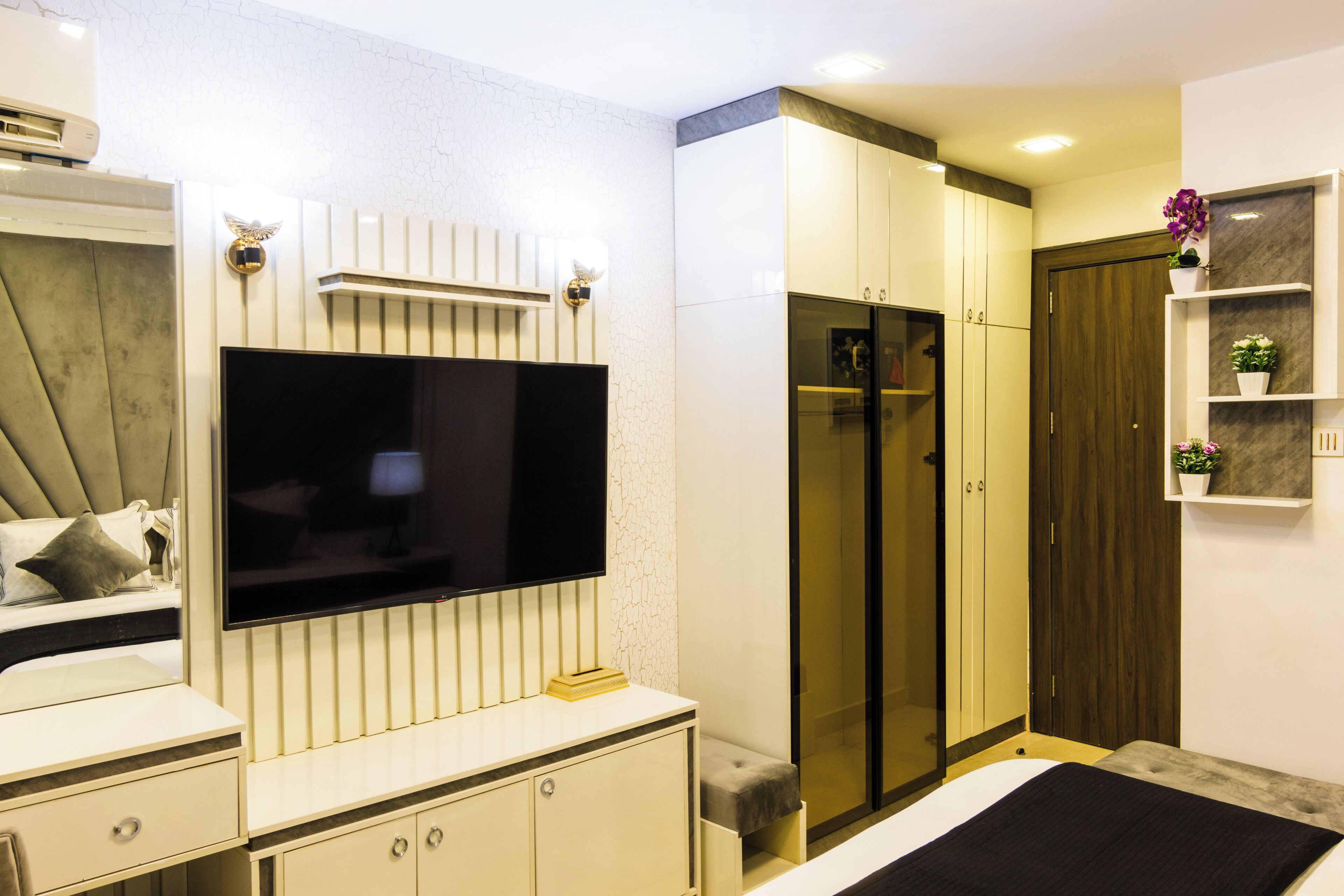 A modern bedroom with a flat-screen TV, a wardrobe, a bedside table, and a built-in shelf with plants.