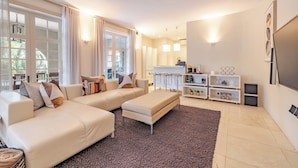 Una sala moderna con un sofá color beige, un otoman a juego y un ventanal con cortinas. Se aprecia un mueble blanco con estantes y un televisor de pantalla plana montado en la pared.