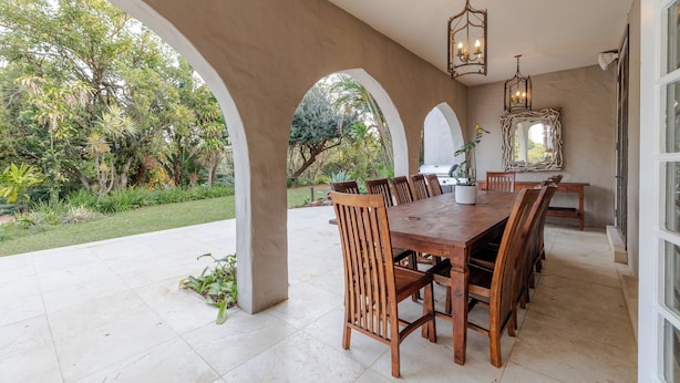 Un área de comedor al aire libre con una larga mesa de madera y sillas, rodeada de arcos y vistas a un jardín exuberante.