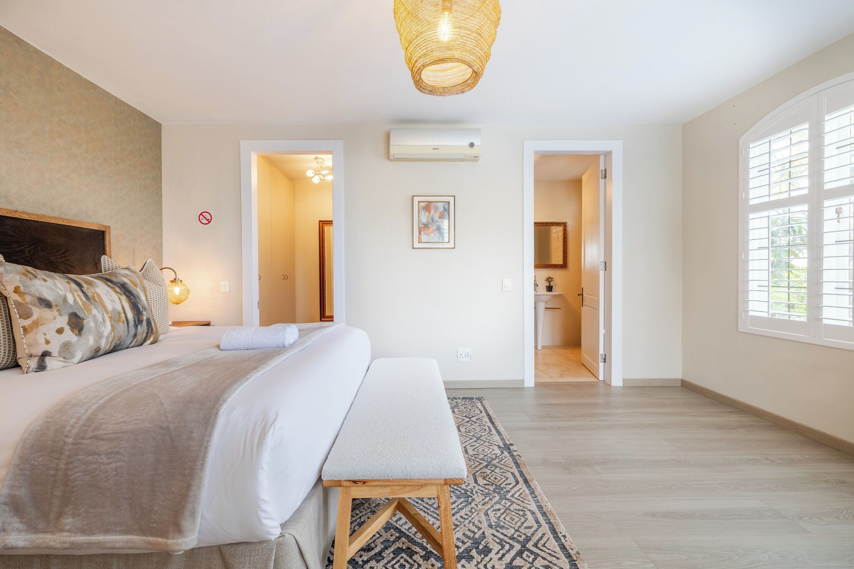 A bedroom with a large bed, a bench, a hanging light fixture, and a door leading to a bathroom.