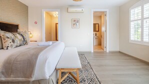 Un dormitorio con una cama grande, un banco, una lámpara colgante y una puerta que da a un baño.