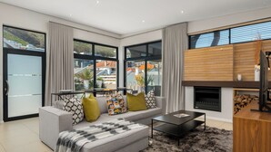 A modern living room with a grey sofa, black coffee table, and a large window with a view of buildings and greenery. - Three Bedroom Family Haven Sea Point (Cape Town)