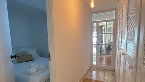 Een smalle gang met een bed in een kamer aan de linkerkant, een houten vloer en een deur naar een balkon.