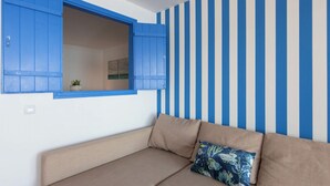 Een kamer met een beige bank, een raam met blauwe luiken en een muur met verticale blauw-witte strepen.