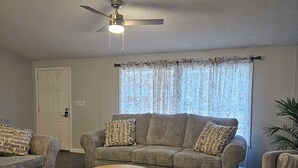 A living room with a grey sofa set, a round coffee table, a ceiling fan, and a window with floral curtains.