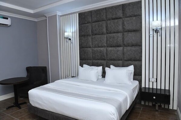 A modern hotel room with a large bed, a chair, a small table, and a wall-mounted air conditioner. - Binos Inn Hotel and Suites  (Benin City)
