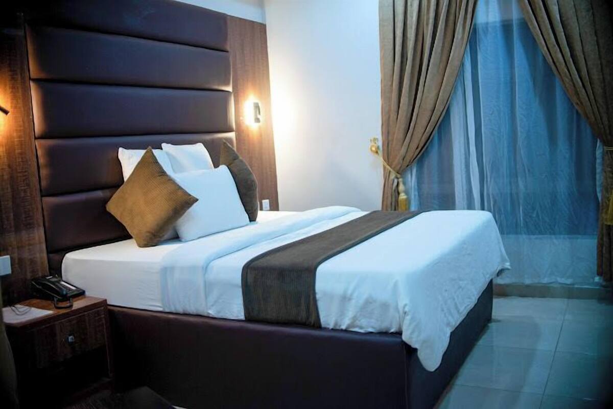 A hotel room with a large bed, a wooden headboard, a bedside table with a phone, and a window with curtains.