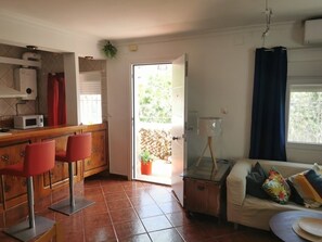 Un espace de vie compact comprenant un coin cuisine, une table à manger avec des chaises rouges, un canapé avec des coussins et, par une porte ouverte, une vue sur un espace extérieur agrémenté d’une plante en pot.