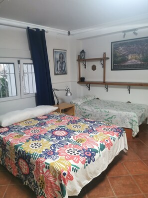 A room with a large bed, a smaller bed, a desk, a chair, a window with curtains, a wall-mounted shelf with decorative items, and a framed picture on the wall.