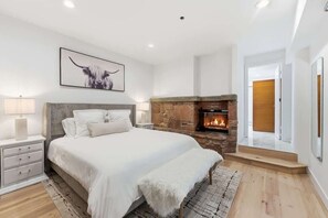 Un dormitorio con chimenea de piedra, una cama con ropa blanca, una mesita de noche y un cuadro en la pared.