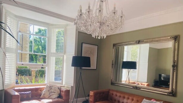 Un salon avec deux canapés, un lustre, un grand miroir et une fenêtre avec des stores.