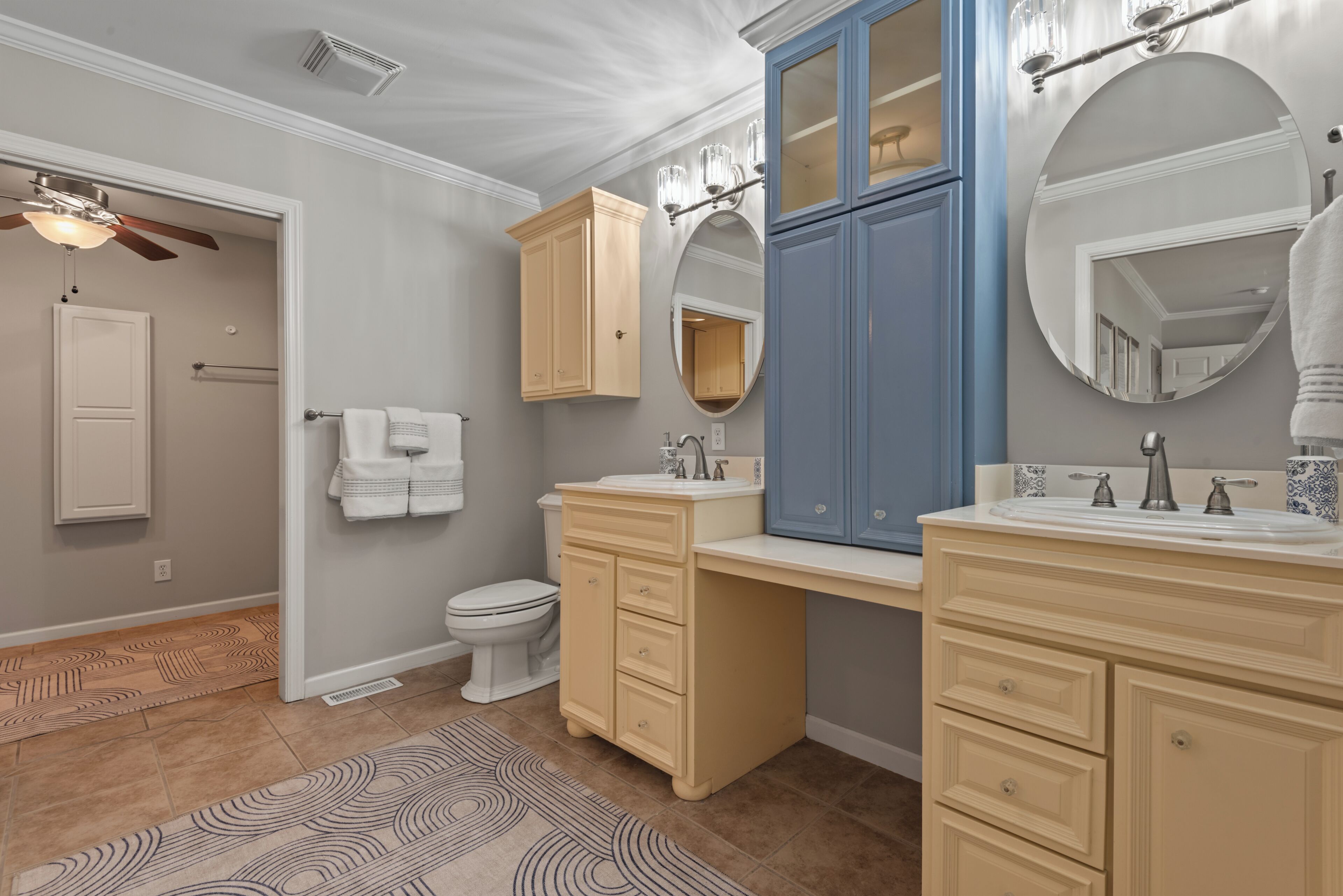 A bathroom with a toilet, two sinks, cabinets, a mirror, and a ceiling fan.