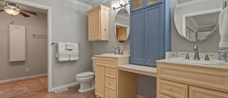 A bathroom with a toilet, two sinks, cabinets, a mirror, and a ceiling fan.