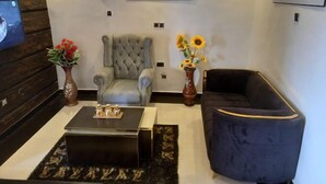 A modern living room with a tufted armchair, a dark blue sofa, a coffee table, and two vases with flowers. - Pefas Ville Hotel  (Benin City)