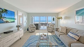 A spacious living room with a large window overlooking the beach, a sofa, a coffee table, and a TV on a wooden cabinet. - Direct Gulf Views Spacious Siesta Key 2 Bed 2 Bath Condo with Views (Siesta Key)