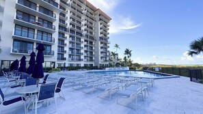 A hotel pool area with lounge chairs and umbrellas, a large building with balconies, and a clear sky. - Tradewinds 214: Beautiful 1 Bedroom Condo on the Beach (No Beach View) (Marco Island)