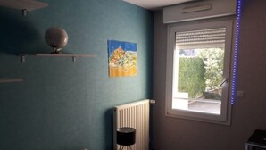 Une chambre avec des murs de couleur turquoise, un lit avec une literie à motifs, un radiateur, une fenêtre avec des stores et un tableau accroché au mur.
