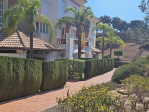 Houses with terracotta tiled roofs line a paved pathway, surrounded by neatly trimmed hedges and small trees.