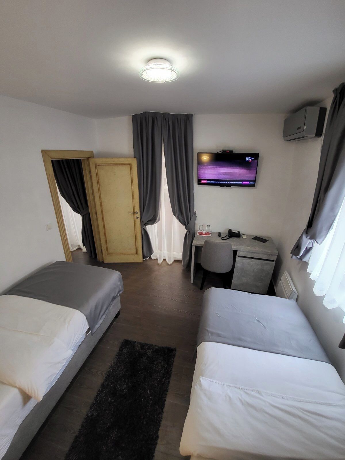 Camera d'albergo con due letti singoli, una scrivania con sedia, una televisione a parete e una porta con tende.