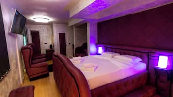 Una habitación con una cama grande, sillas tapizadas en rojo, un televisor de pantalla plana e iluminación ambiental morada.