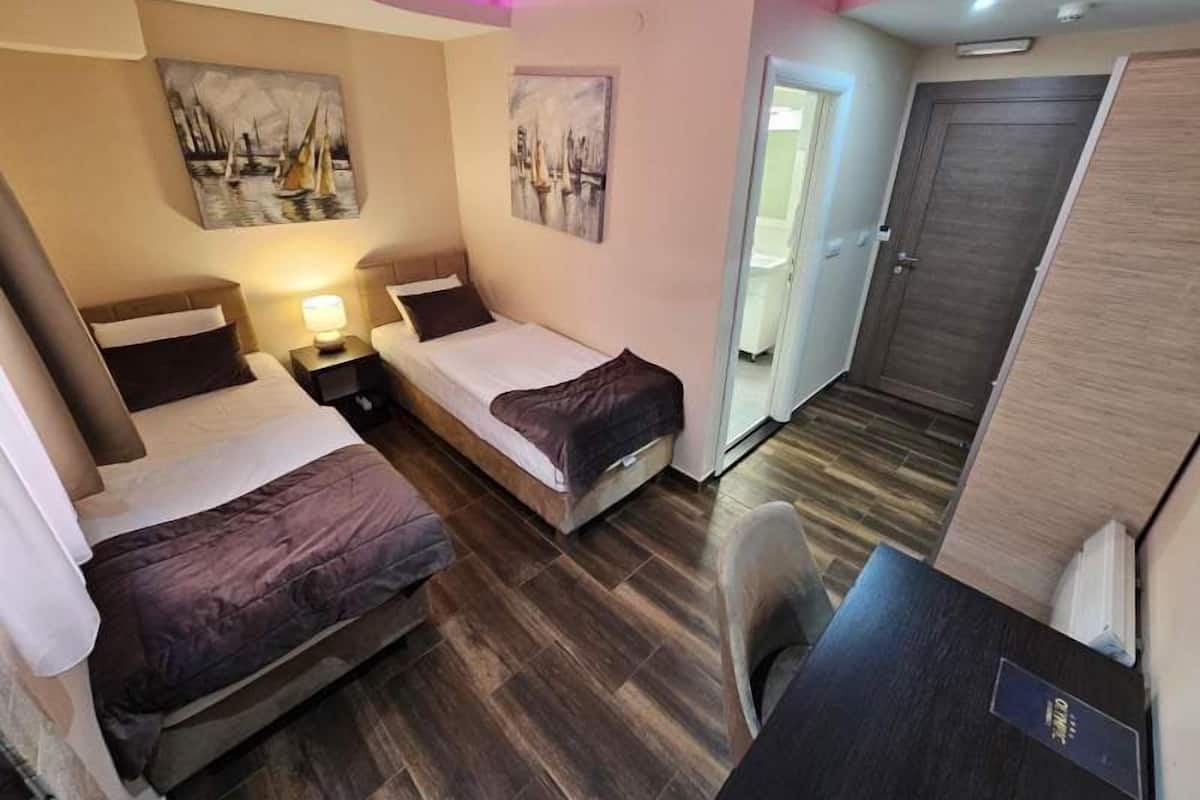 Hotellihuone, jossa on kaksi erillistä sänkyä, työpöytä ja tuoli. Seinillä on tauluja, ja oikealla puolella on ovi.
