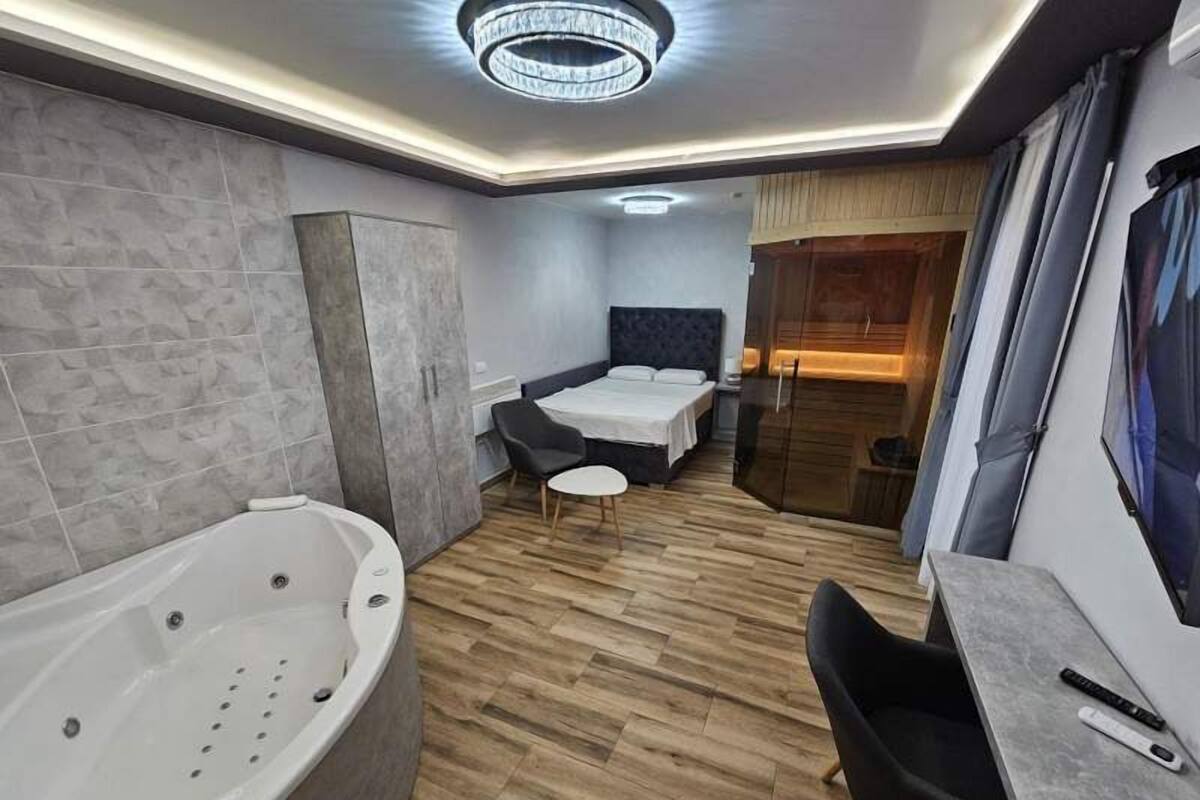 Moderni hotellihuone, jossa on suuri kylpyamme, tummanvärinen päätyosalla varustettu sänky, pieni pyöreä pöytä ja työpöytä tuolilla.