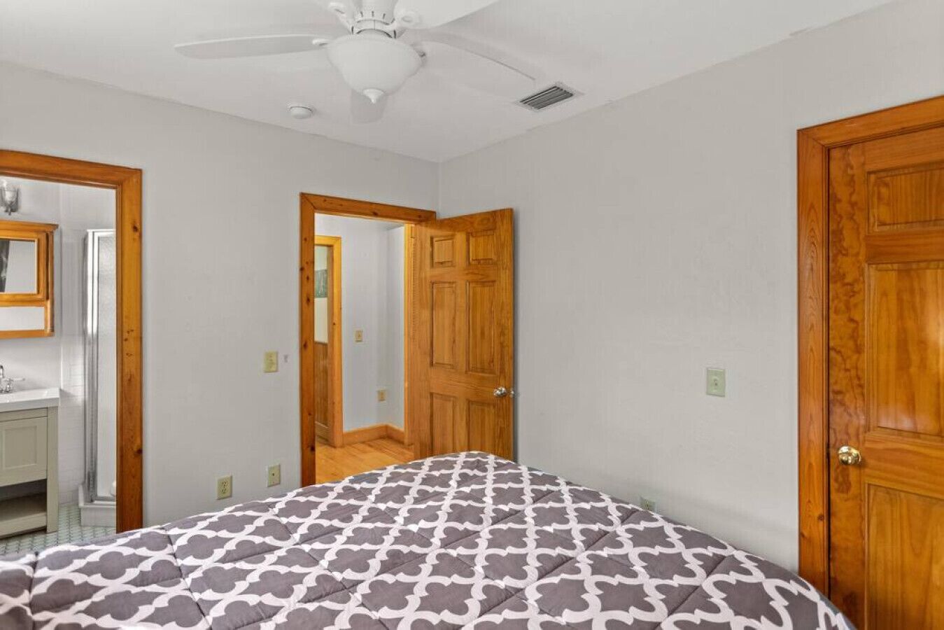 Un dormitorio con una cama, ventilador de techo, puertas de madera y un baño visible a través de un vano.