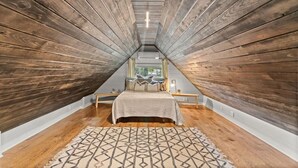 Une chambre avec un plafond en pente, des murs en bois, un lit surmonté d’un tapis à motifs et une fenêtre avec des rideaux.