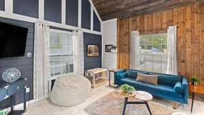 Un salon avec un canapé bleu, un pouf blanc rond, une petite table en bois sur laquelle est posée une plante, et un téléviseur sur un meuble noir.