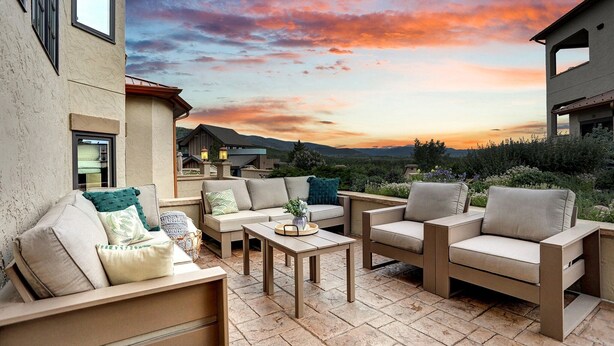 Une terrasse extérieure avec des canapés et des fauteuils de couleur beige, une table en bois et une vue sur un coucher de soleil se déclinant sur un paysage montagneux.