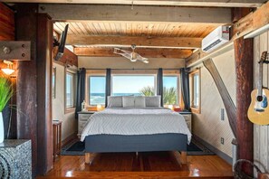 Een slaapkamer met een groot bed, houten balken in het plafond, uitzicht op de oceaan en een gitaar aan de muur.