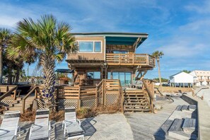 A beachfront house with a wooden deck, outdoor seating, and a large window overlooking the ocean.