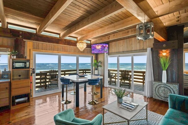 A spacious room with a wooden ceiling, large windows offering ocean views, a dining area with blue chairs, and a TV mounted on the wall.
