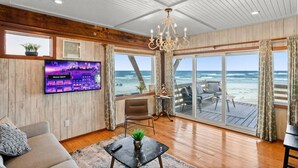 A spacious living room with a large TV, wooden walls, a chandelier, and a view of the ocean through large sliding doors. - Treehouse Artist Haven Direct Oceanfront 2br (St. Augustine)