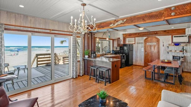 A spacious kitchen with a large island, a dining area with a wooden table and chairs, and a view of the beach through large sliding glass doors. - Treehouse Artist Haven Direct Oceanfront 2br (St. Augustine)