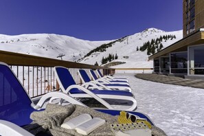 Fileira de cadeiras de descanso azuis com um livro e óculos de sol em um banco de pedra, com vista para uma montanha nevada e um edifício com amplas janelas.